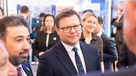Menschen in formeller Kleidung stehen auf einer Messe dicht beieinander im Gespräch im Vordergrund ein Mann im Anzug mit Krawatte