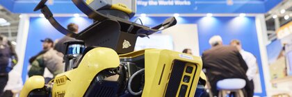 Gelber Roboterhund auf dessen Rücken ein Gerät montiert ist, vor einem Messestand mit Menschen im Hintergrund.