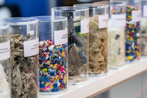Aufgereihte transparente Zylinder, die beschriftet und mit unterschiedlichen Recyclingmaterialien befüllt sind.