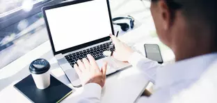 A businessman sits in front of an open laptop and types. To the left of the laptop is a to-go coffee cup on top of a notebook.