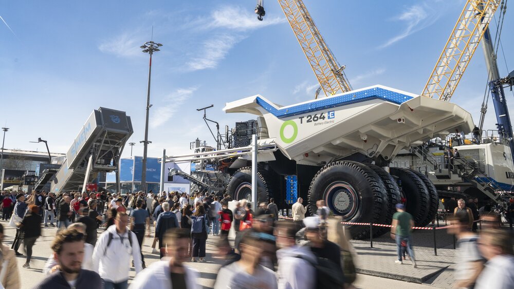 Menschenmenge auf einer Messe vor einem riesigen Muldenkipper und Baumaschinen unter blauem Himmel.