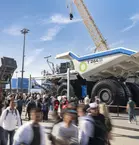 Crowd at a trade fair in front of a huge dump truck and construction machinery under a blue sky.