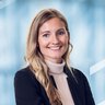 Lächelnde Frau mit langen blonden Haaren in schwarzem Blazer und beigem Rollkragenpullover, vor einem unscharfen blauen Hintergrund.
