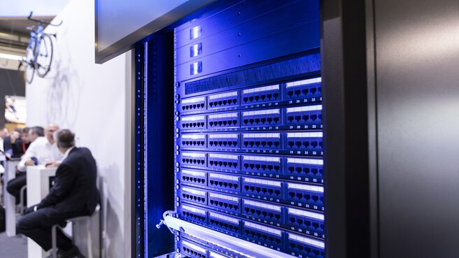 Blue illuminated server rack with numerous connection sockets on the wall of an exhibition stand.