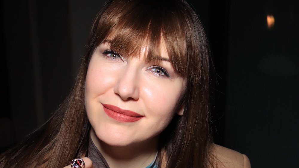 A smiling woman with long brown hair is wearing a designer top and a brown blazer, an eye-catching brooch and a ring on her finger.