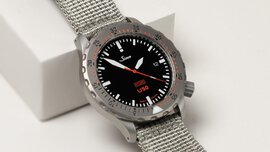 A close-up view of a wristwatch with a black dial, minute markers on the outer edge, and a fabric strap.