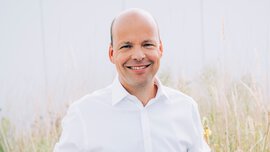 Ein glatzköpfiger Mann in einem weißen Hemd lächelt, während er im Freien zwischen hohem Gras und einem Betonvorsprung steht.