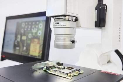 Nahaufnahme eines Lichtmikroskops, unter dem eine offene Platine liegt. Im Hintergrund ist ein Monitor zu sehen.
