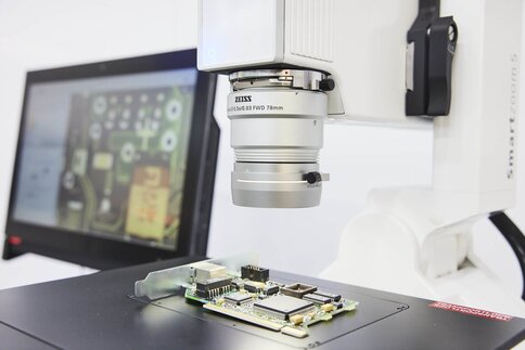 Close-up of an optical microscope with an open circuit board underneath. A monitor can be seen in the background.