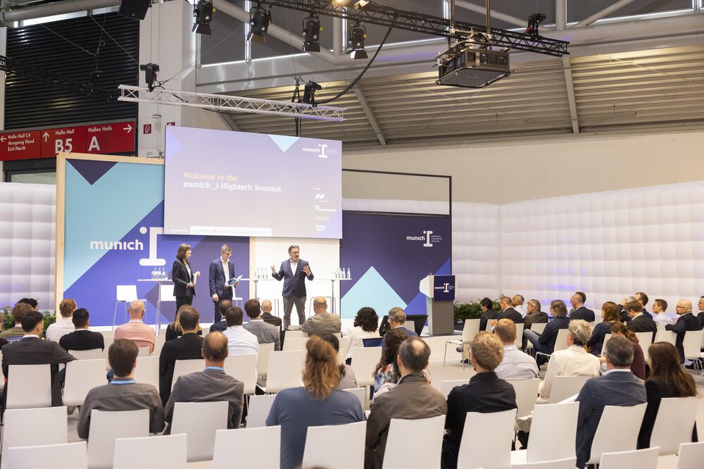 A woman and two men in business attire stand on a stage at the ‘munich i’ and give a presentation to an interested audience.