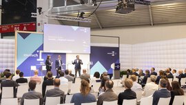 A woman and two men in business attire stand on a stage at the ‘munich i’ and give a presentation to an interested audience.