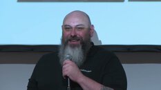 Ein Mann mit Bart spricht vor einem Bildschirm in ein Mikrofon.