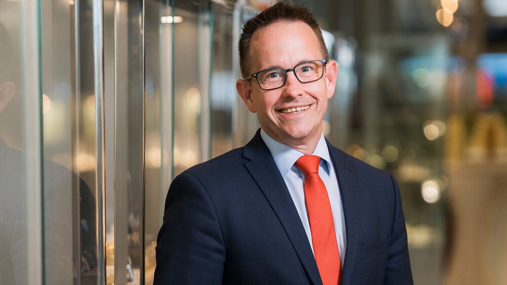 A smiling man with short brown hair and glasses is wearing a dark blue jacket and a red tie. He is standing next to a reflective display case.
