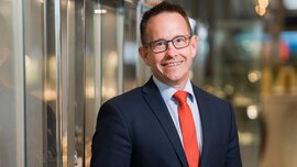 A smiling man with short brown hair and glasses is wearing a dark blue jacket and a red tie. He is standing next to a reflective display case.