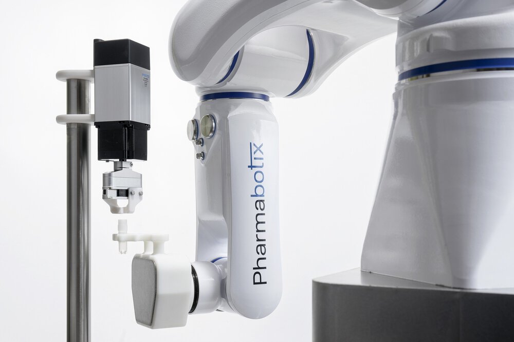 Roboterarm mit der Aufschrift „Pharmabotix“ und einem Greifwerkzeug, positioniert neben einem kleinen zylindrischen Gerät.