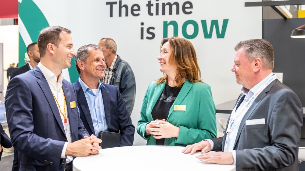 Three older men in suits and a brown-haired woman in a green jacket are standing in a semi-circle talking. In the background is a billboard in white and green with the inscription The time is now.