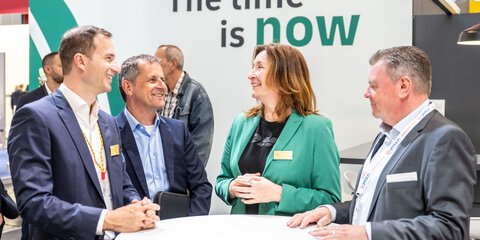Three older men in suits and a brown-haired woman in a green jacket are standing in a semi-circle talking. In the background is a billboard in white and green with the inscription The time is now.