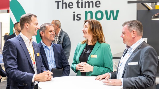 Three older men in suits and a brown-haired woman in a green jacket are standing in a semi-circle talking. In the background is a billboard in white and green with the inscription The time is now.
