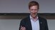 A smiling speaker with a headset gestures during a presentation.