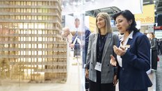 Two women are looking at a detailed architectural model of a modern building at EXPO REAL in Munich. One woman is explaining something and gesturing with her hands, while the other listens attentively and smiles. In the background, other trade fair visitors and exhibitor booths can be seen, including one with the sign “Savills.”