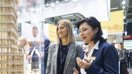 Zwei Frauen betrachten auf der EXPO REAL in München ein detailliertes Architekturmodell eines modernen Gebäudes. Eine der Frauen erklärt etwas und gestikuliert dabei, während die andere aufmerksam zuhört und lächelt. Im Hintergrund sind weitere Messebesucher und Ausstellerstände zu sehen, darunter ein Stand mit dem Schriftzug „Savills“