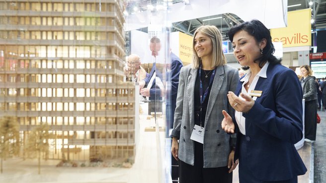 Zwei Frauen betrachten auf der EXPO REAL in München ein detailliertes Architekturmodell eines modernen Gebäudes. Eine der Frauen erklärt etwas und gestikuliert dabei, während die andere aufmerksam zuhört und lächelt. Im Hintergrund sind weitere Messebesucher und Ausstellerstände zu sehen, darunter ein Stand mit dem Schriftzug „Savills“