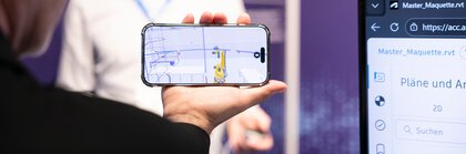 Person im Vordergrund hält ein Smartphone in der Hand, auf dem ein technischer Zeichenplan zu sehen ist. Im Hintergrund ist eine Person mit VR-Brille zu sehen.