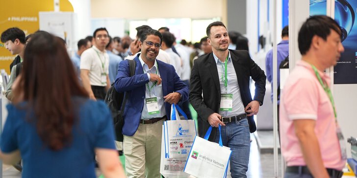 Two men in business attire walk through a busy convention hall, smiling and carrying event bags.