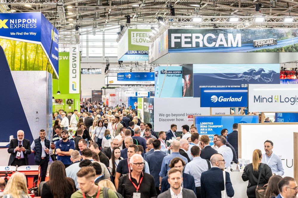 Eine Menschenmenge, die durch eine belebte Logistikmesse mit Ständen und Bannern verschiedener Unternehmen läuft.