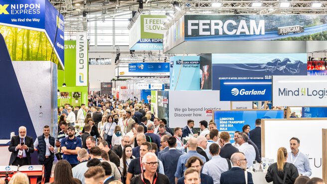 Eine Menschenmenge, die durch eine belebte Logistikmesse mit Ständen und Bannern verschiedener Unternehmen läuft.