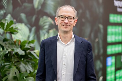 Portrait of a smiling man wearing glasses, a dark blue blazer and white shirt, standing in front of a blurred leafy exhibition backdrop with large green plants behind him.