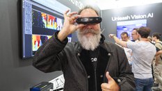 Ein Mann mit grauem Vollbart hält eine 3D-Kamera vor seine Augen und zeigt mit der anderen Hand den Daumen nach oben. Im Hintergrund ist ein Messestand mit Bildschirmen und dem Schriftzug „Unleash Vision AI“ zu sehen.