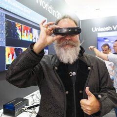 A man with a gray beard  holds a 3D camera in front of his eyes and gives a thumbs-up with his other hand. In the background, a trade show booth with screens and the slogan “Unleash Vision AI” is visible.
