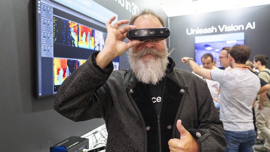 Ein Mann mit grauem Vollbart hält eine 3D-Kamera vor seine Augen und zeigt mit der anderen Hand den Daumen nach oben. Im Hintergrund ist ein Messestand mit Bildschirmen und dem Schriftzug „Unleash Vision AI“ zu sehen.