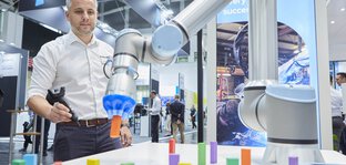 A man uses a remote control to operate a robotic arm that manipulates colorful dominoes at an exhibition stand.