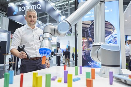 A man uses a remote control to operate a robotic arm that manipulates colorful dominoes at an exhibition stand.