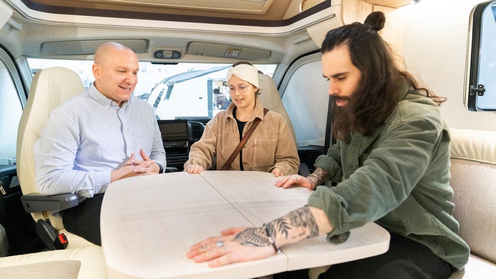 Drei Personen, die in einem Wohnmobil sitzen, sich um einen Tisch herum unterhalten und dessen Oberfläche untersuchen.