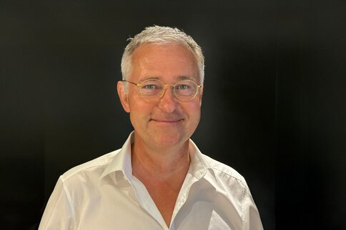 Smiling man with short gray hair and glasses, wearing a white shirt and standing in front of a black background.