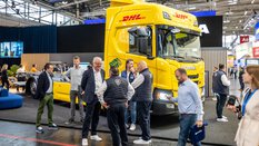 Mehrere Messebesucher stehen vor einem gelben DHL-Truck ohne Ladung und unterhalten sich. Im Hintergrund sieht man in die Halle A6.