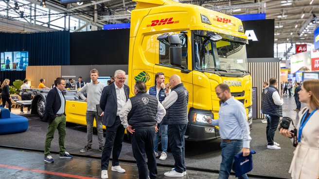 Several trade fair visitors are standing in front of a yellow DHL truck without a load and chatting. Hall A6 can be seen in the background.