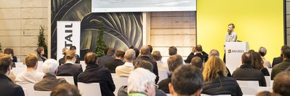 Ein Mann hält während der BAU-Messe vor einem aufmerksamen sitzenden Publikum einen Vortrag.