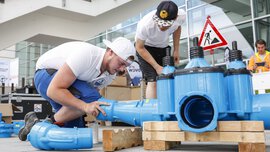 Two workers install a complex pipe system as part of a demonstration in front of an exhibition hall.