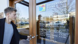 Junger Mann öffnet eine Glastür zu einem Gebäude, auf der das Logo des Bauzentrum Poing und dessen Öffnungszeiten stehen.