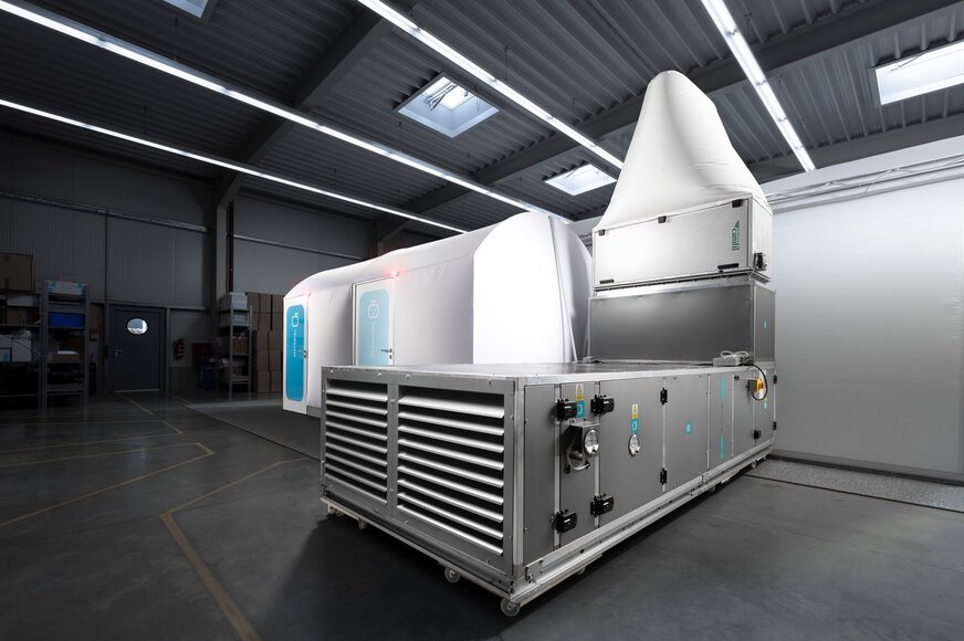 A white mobile clean room with two doors stands in a hall next to a large filter system.
