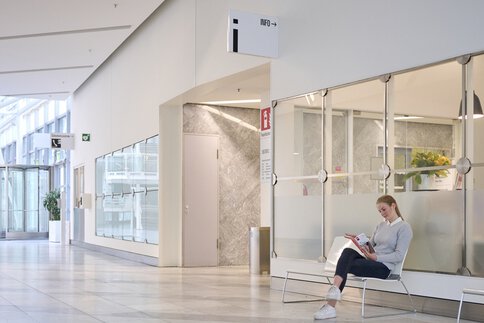 Das Foyer des MOC Ordercenters mit Blick auf die verglasten Außenwände des Infocenters. Eine Frau sitzt auf einer Bank davor und liest.