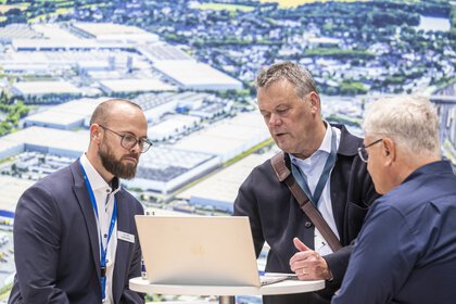 Drei Männer stehen an einem runden Stehtisch und schauen konzentriert auf einen Laptop. Im Hintergrund ist ein großes Luftbild eines Industrie- und Logistikareals zu sehen.