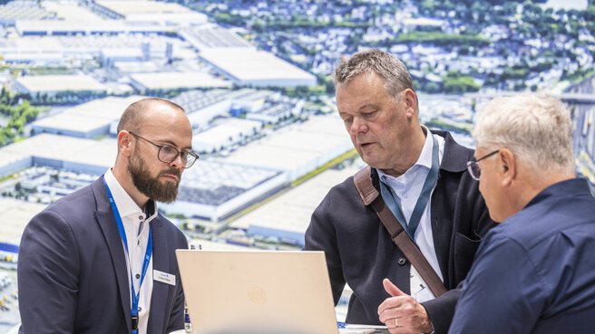 Drei Männer stehen an einem runden Stehtisch und schauen konzentriert auf einen Laptop. Im Hintergrund ist ein großes Luftbild eines Industrie- und Logistikareals zu sehen.