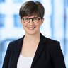 A smiling woman with round glasses, short dark hair and a dark blazer.