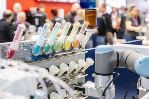 An industrial robot arm works on a machine with several tubes filled with colorful liquids. In the background, people at a trade fair are blurred.