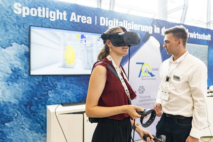 A young woman operates a presentation with VR glasses at a trade fair stand, her progress is visible on a screen in the background.